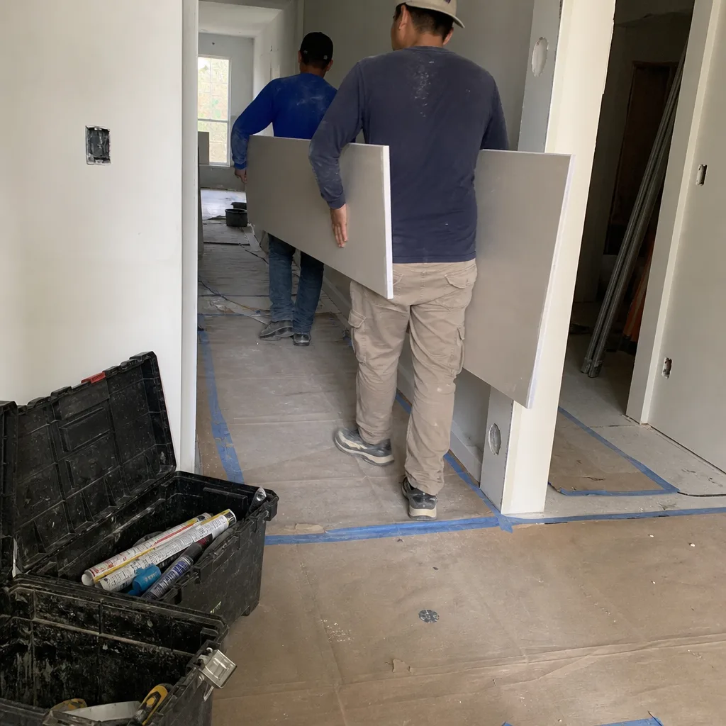 Two construction workers carrying a large drywall panel during home renovation.